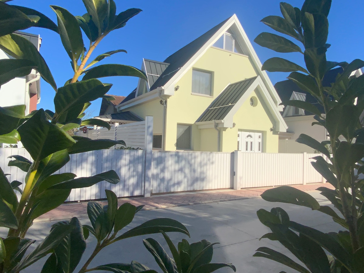 The exterior of a modern, detached house is visible, showcasing a light yellow color with distinctive rooflines. A white fence and well-maintained plants frame the property, while a clear blue sky is seen overhead.