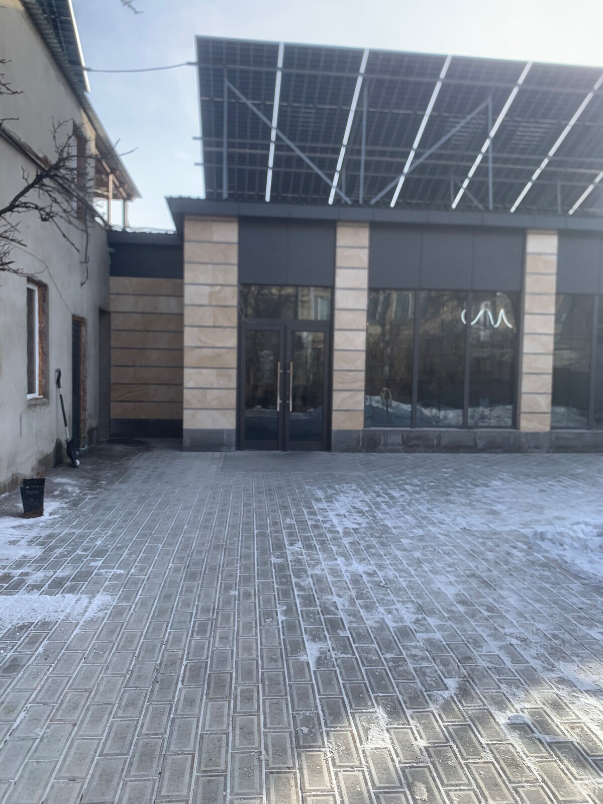 The exterior of a modern building is featured, showcasing large glass windows and a stone façade. Solar panels are positioned on the roof, providing a contemporary touch. A paved area in front is partly covered with a light dusting of snow, indicating a cold climate.