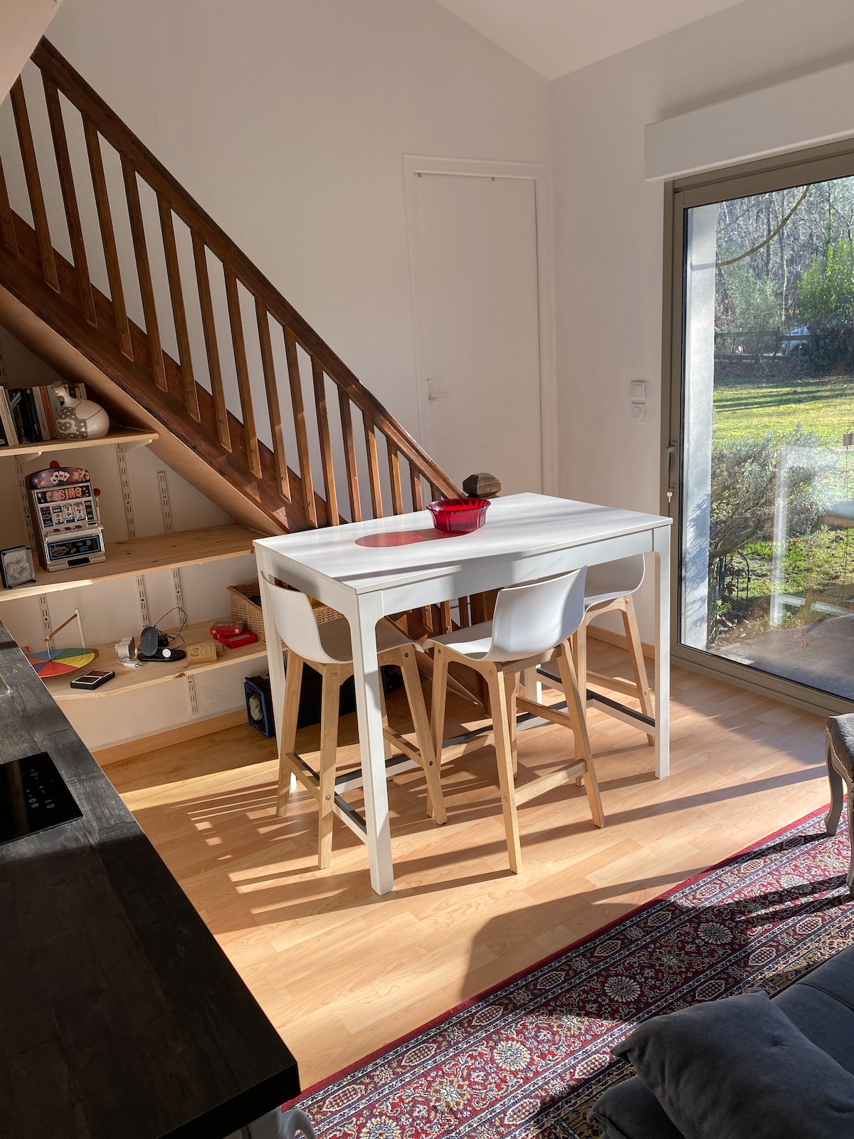 A dining area features a white table accompanied by two light wooden chairs. Natural light streams through a sliding glass door, illuminating the space. A staircase is visible in the background, along with a decorative shelf positioned nearby.
