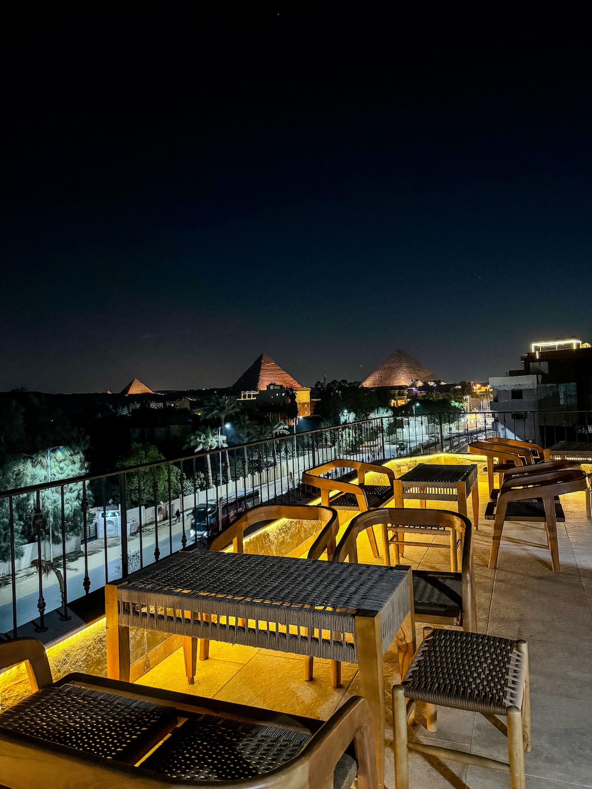 A rooftop terrace is illuminated at night, featuring wooden tables and chairs arranged along a railing. The distant Pyramids can be seen in the background, surrounded by a softly lit landscape. The setting offers a unique view of the night sky and nearby structures.