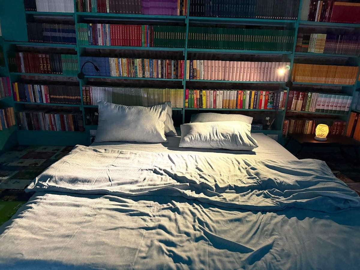 A spacious bed is situated in a cozy nook, surrounded by shelves filled with various books. Soft bedding provides comfort, while a warm lamp emits a gentle glow. The backdrop is a vibrant display of colorful book spines, creating an inviting reading environment.