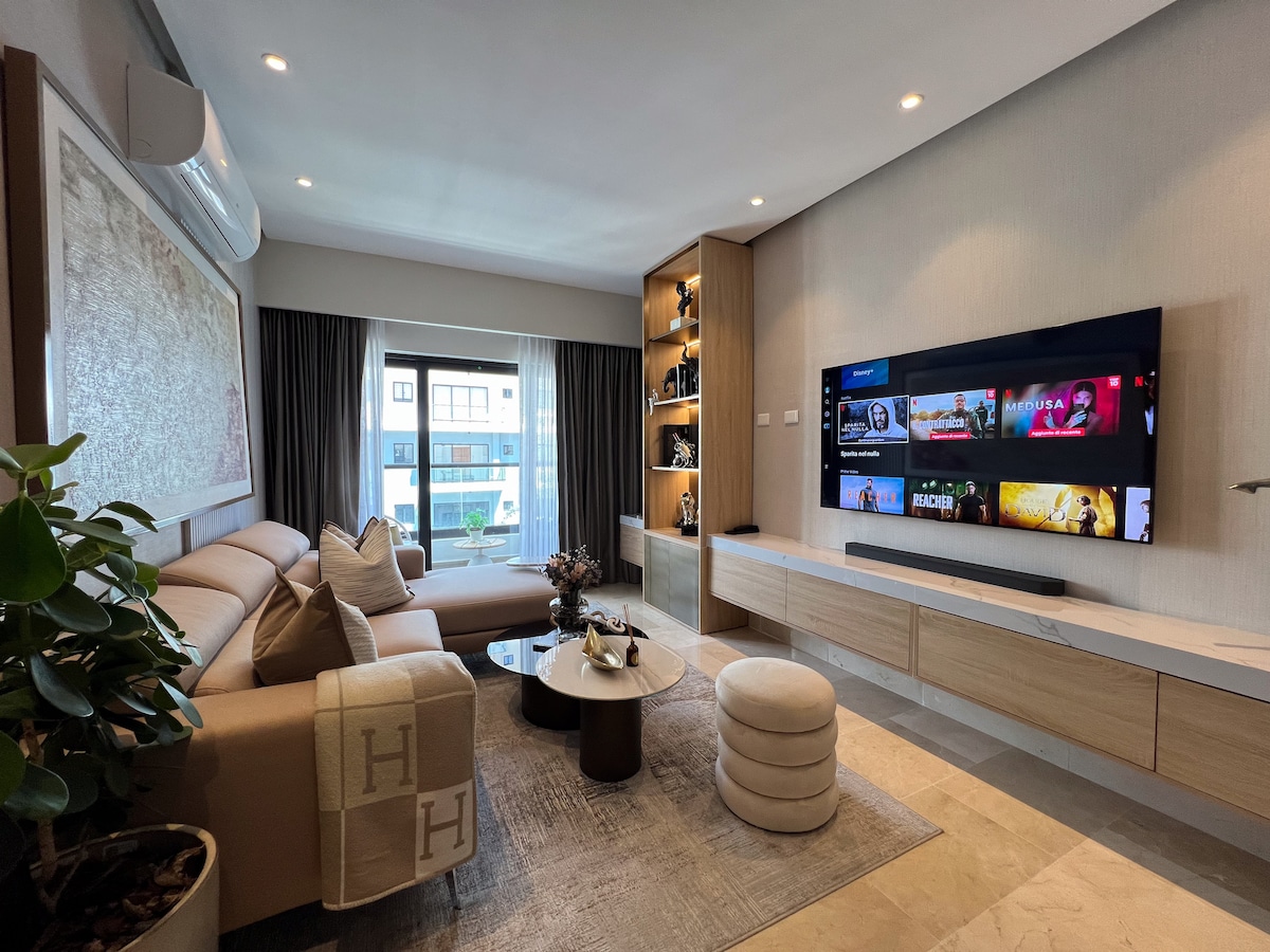 A modern living room features a plush sectional sofa paired with a round coffee table. Natural light enters through the large window, adorned with dark curtains. A wall-mounted television displays various content, while decorative shelves showcase curated items, contributing to a comfortable atmosphere.
