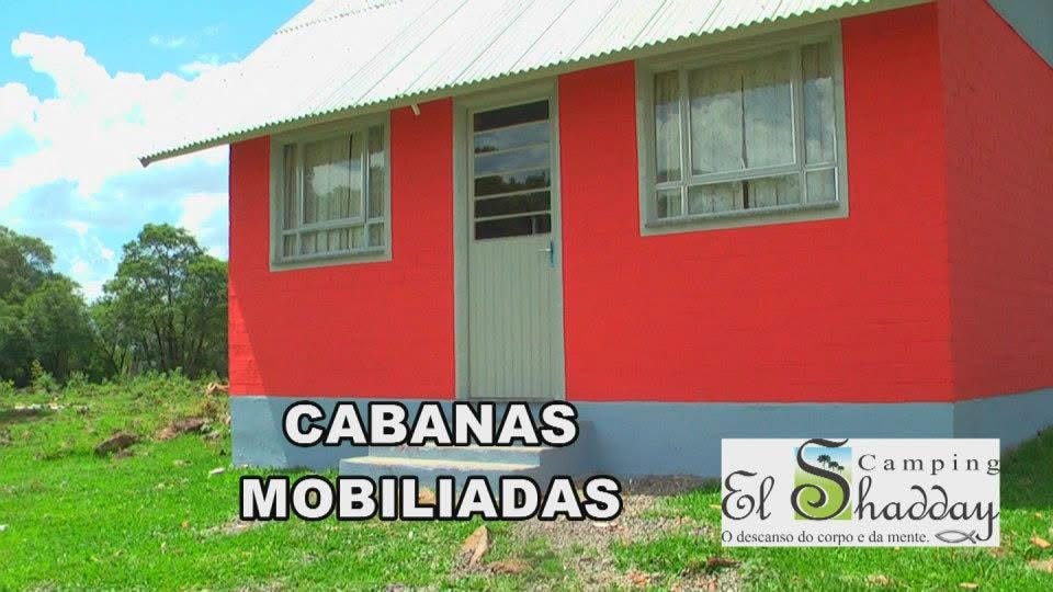 A small, bright red cabin is featured, with two rectangular windows on either side of a central door. The building is set in a grassy area, surrounded by greenery, providing a welcoming appearance for outdoor enthusiasts.