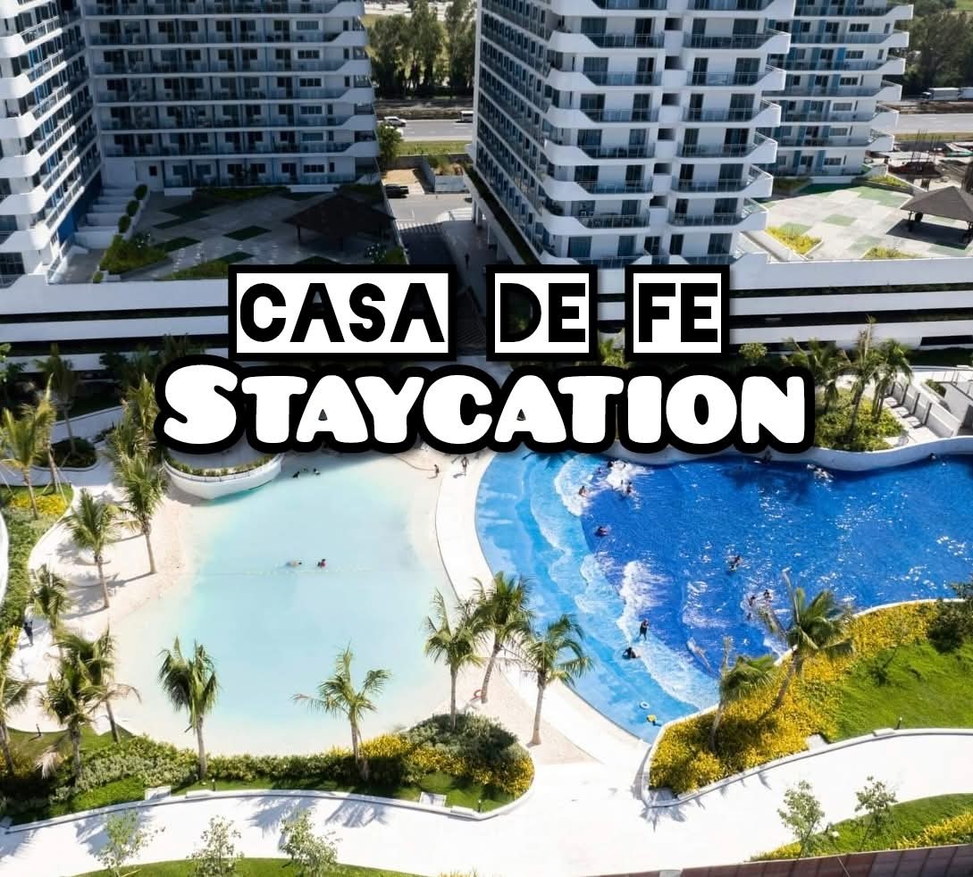 An aerial view showcases a wave pool alongside a tranquil beach area, surrounded by lush greenery and palm trees. The modern buildings in the background provide a contemporary setting. The two pools are distinct, with one featuring gentle waves and the other a calm, inviting surface.