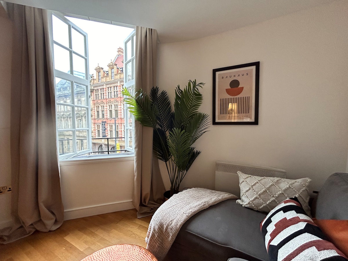 A spacious lounge area features large windows with dual-opening panes, allowing natural light to fill the room. A cozy gray sofa is adorned with decorative pillows, and a leafy indoor plant adds a touch of greenery. City views are visible through the windows, showcasing the nearby architecture.
