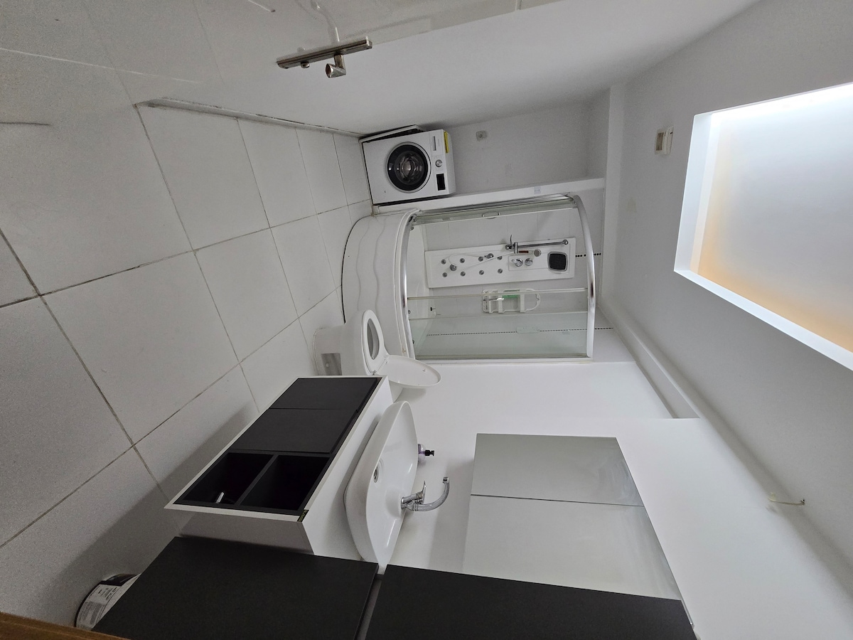 A clean and modern bathroom is presented, featuring a compact shower with glass doors. A white toilet and sink are positioned against the wall, with a small cabin for storage. A washing machine occupies one corner, contributing to the overall functionality of the space.