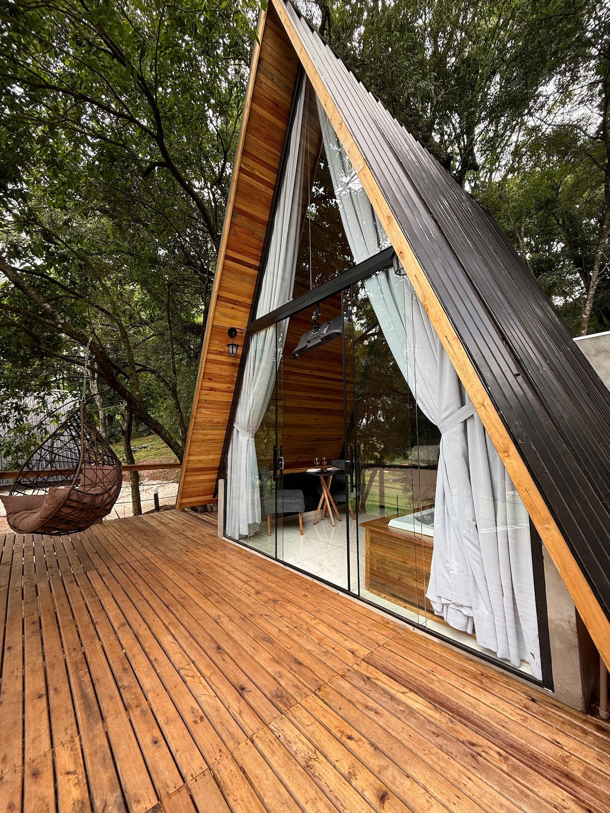 A modern A-frame cabin is surrounded by trees, featuring large glass windows that allow natural light to fill the space. A wooden deck leads to a cozy seating area with a hanging chair, inviting relaxation in a serene outdoor setting.