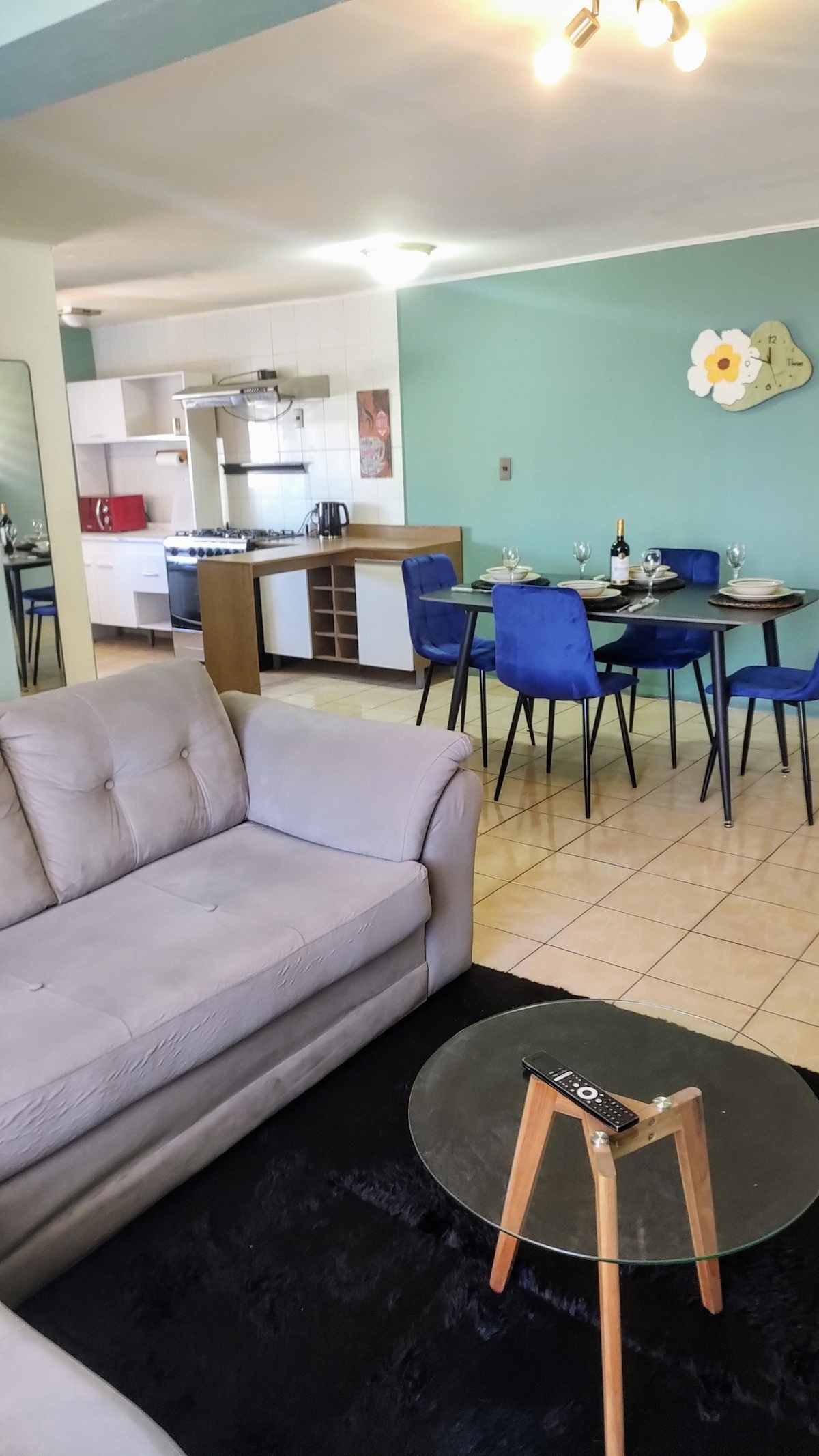 A comfortable living area features a light-colored sofa positioned near a small round table with a wooden leg. The open kitchen and dining area are visible in the background, showcasing a dining set with blue chairs and a well-equipped kitchen counter.