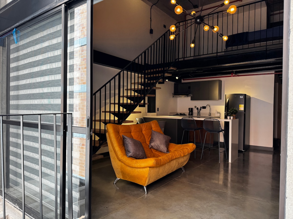 An open-concept space is showcased, featuring a modern kitchen and a cozy, amber-colored sofa. Industrial-style lighting complements the ambiance. A flight of stairs leads to an upper level, while large glass doors provide a seamless connection to the balcony.
