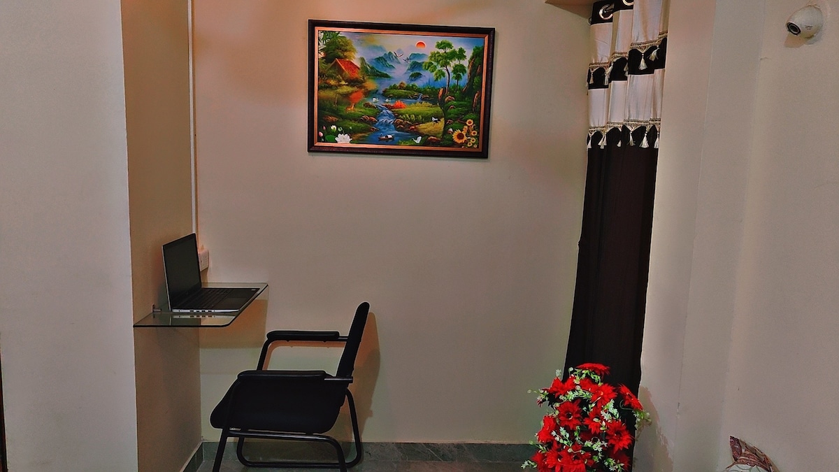 A small corner features a glass desk paired with a black chair, illuminated by soft lighting. A colorful landscape painting adorns the wall above. Decorative flowers in a vase provide a touch of vibrancy in the otherwise neutral space.