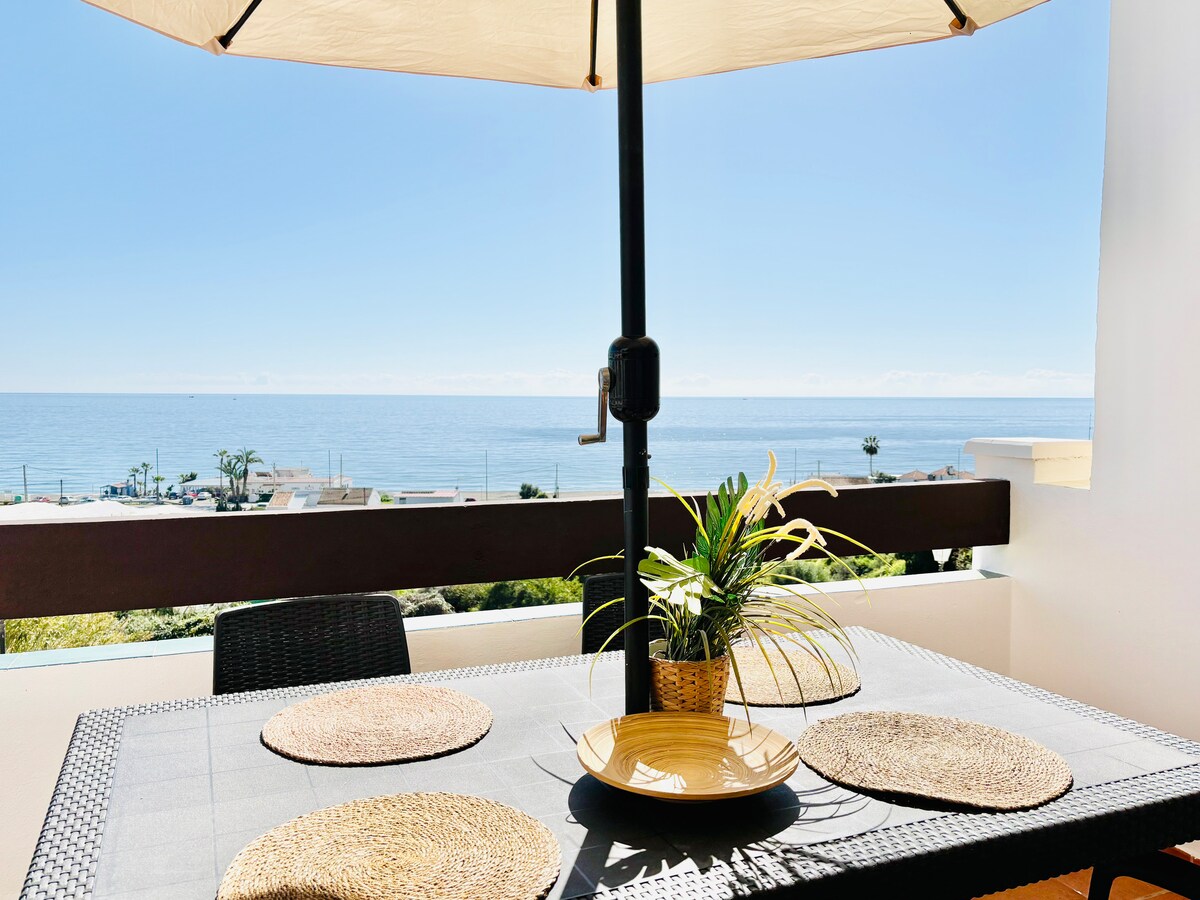 A dining table is positioned on a terrace, featuring a large umbrella providing shade. Four placemats are arranged on the table, which is adorned with a potted plant. The ocean is visible in the background under a clear blue sky.