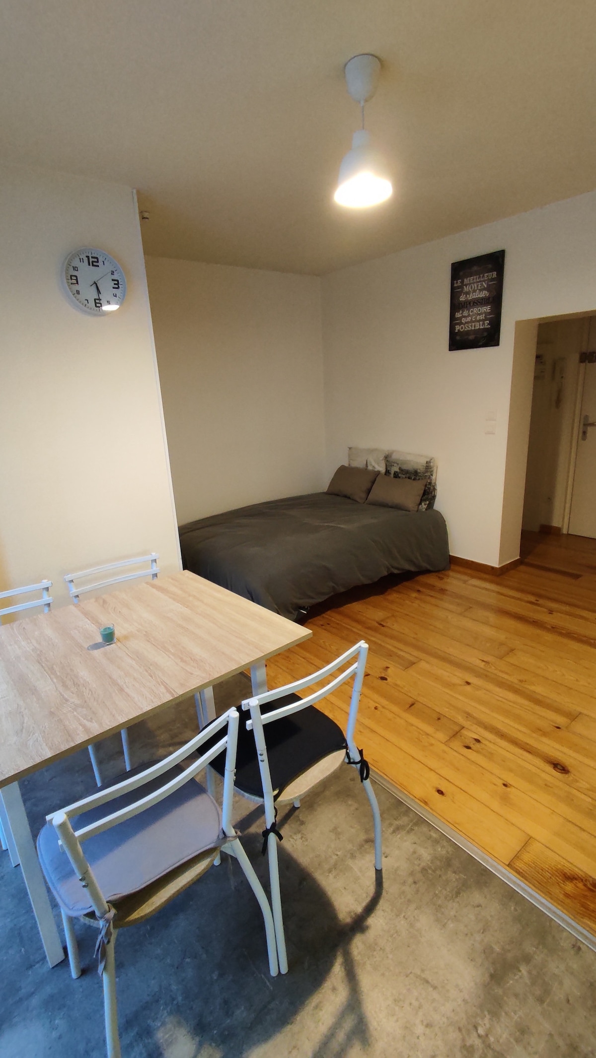 A modern studio layout is presented, featuring a spacious bed with neutral bedding and a dining area with a wooden table complemented by two chairs. Natural light illuminates the light-colored walls and polished wooden flooring, creating a welcoming ambiance. A clock adorns one wall.