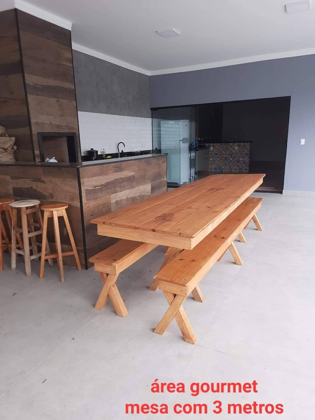 A spacious gourmet area features a long wooden table measuring three meters, surrounded by benches on either side. A kitchen countertop with a dark finish is visible in the background, complemented by wooden stools. The flooring is light-colored, contributing to an open and airy feel.