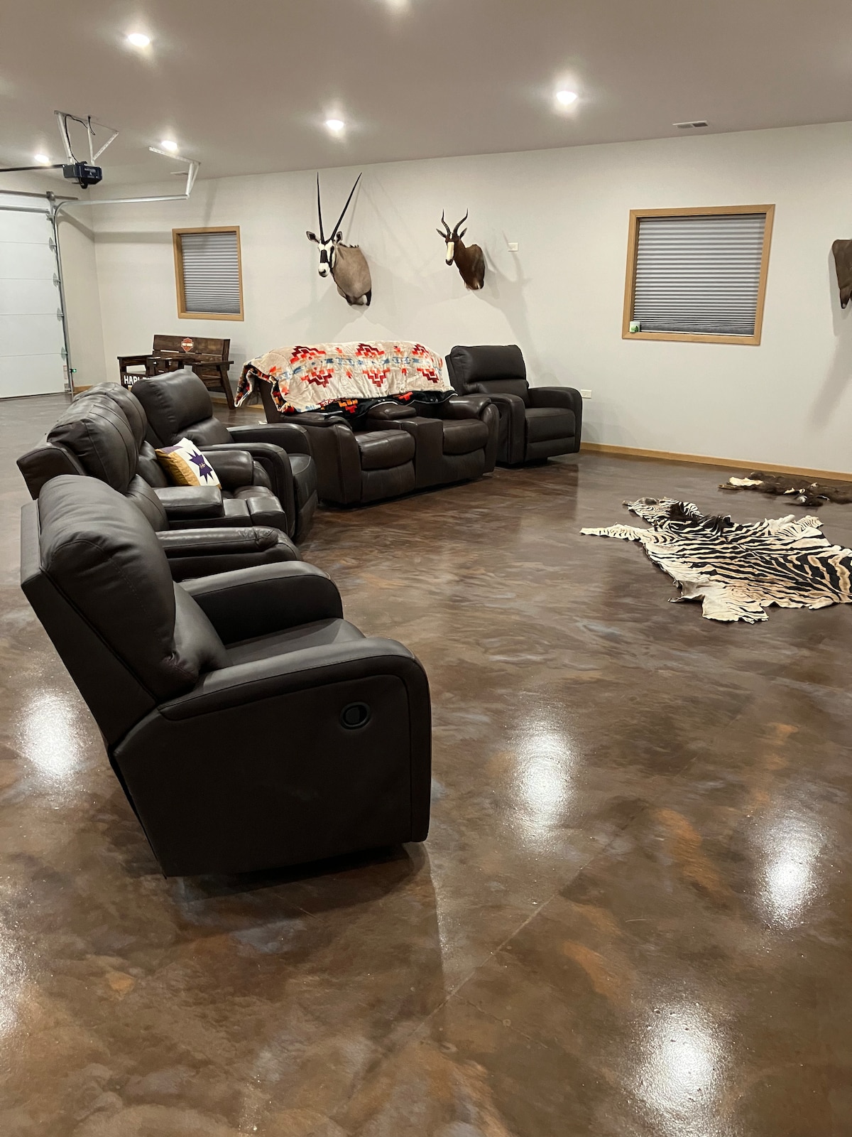 A spacious social area features reclining leather chairs arranged for comfort. A patterned throw covers a couch, while a large animal hide rug is placed on a polished concrete floor. Natural light enters through windows with horizontal blinds.