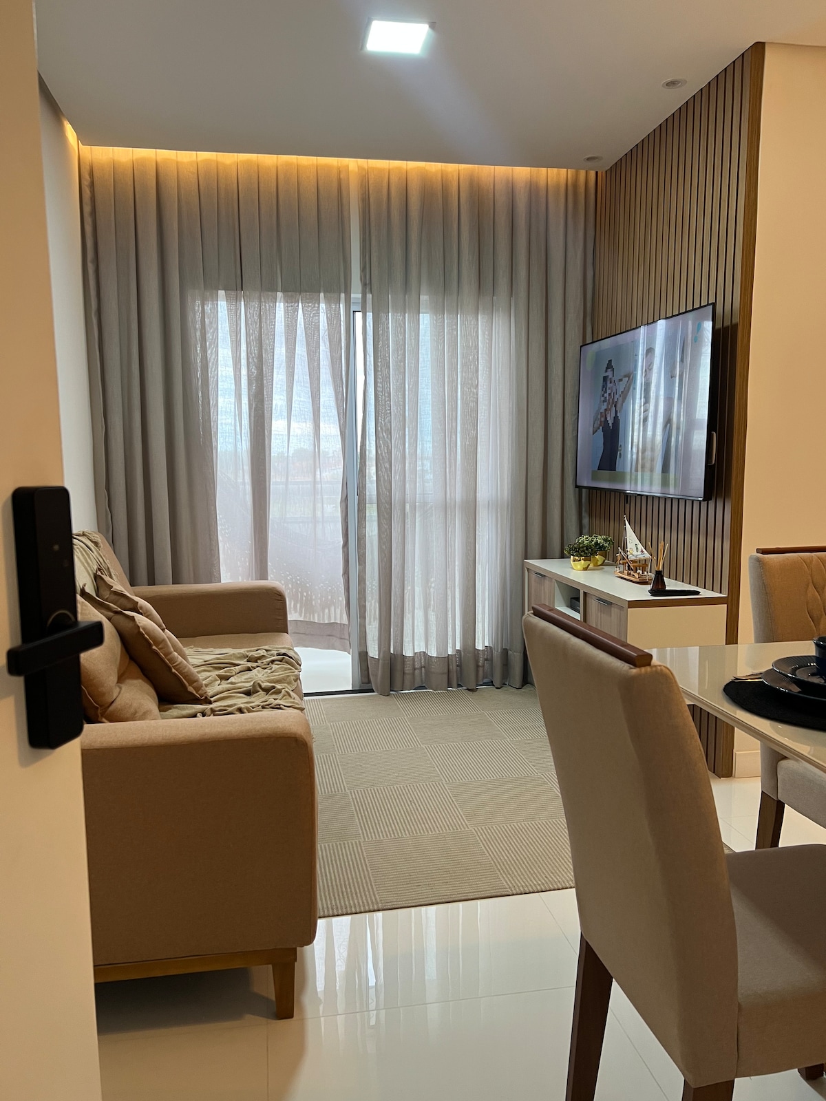 A cozy living area features a soft sofa positioned near large, sheer curtains that softly filter light. A wall-mounted television is visible, and a dining table is elegantly set, enhancing the inviting atmosphere of the space.