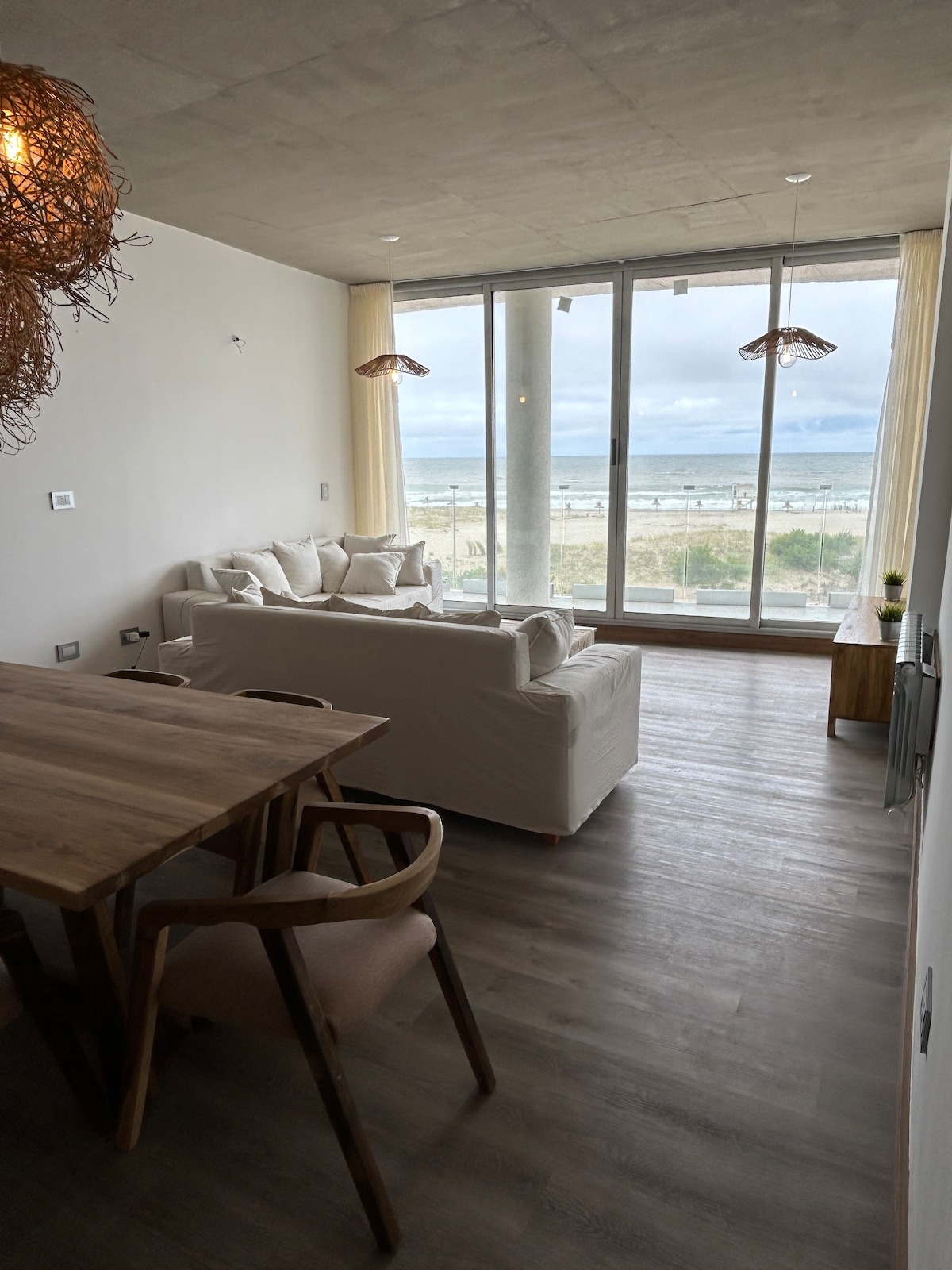 A bright living area features a light-colored sofa with ample seating and a wooden dining table nearby. Large glass windows offer a clear view of the ocean and bring natural light into the space, creating an open and airy environment.