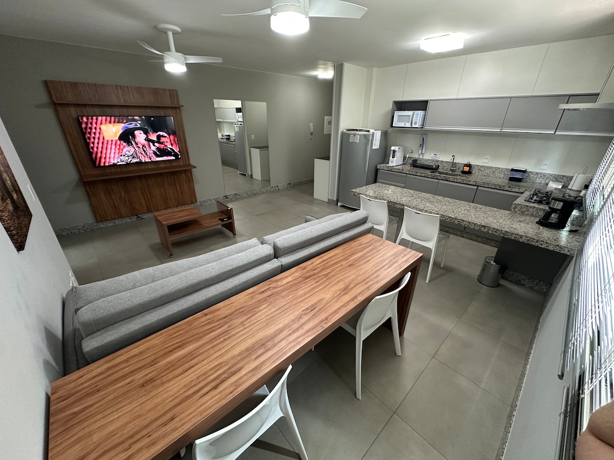 A modern open-concept living area features a spacious gray sofa facing a large flat-screen television. A wooden dining table is set for six, surrounded by white chairs. The kitchen, equipped with contemporary appliances, is visible in the background, highlighting the sleek design.