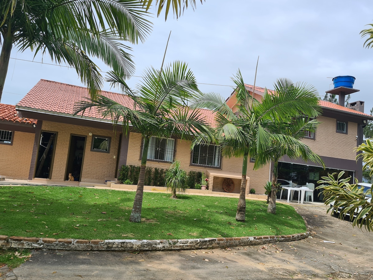 A spacious property is surrounded by lush green grass and palm trees, featuring a welcoming façade with a tiled roof. The exterior includes multiple entrances and a parking area, with outdoor seating visible in the background.