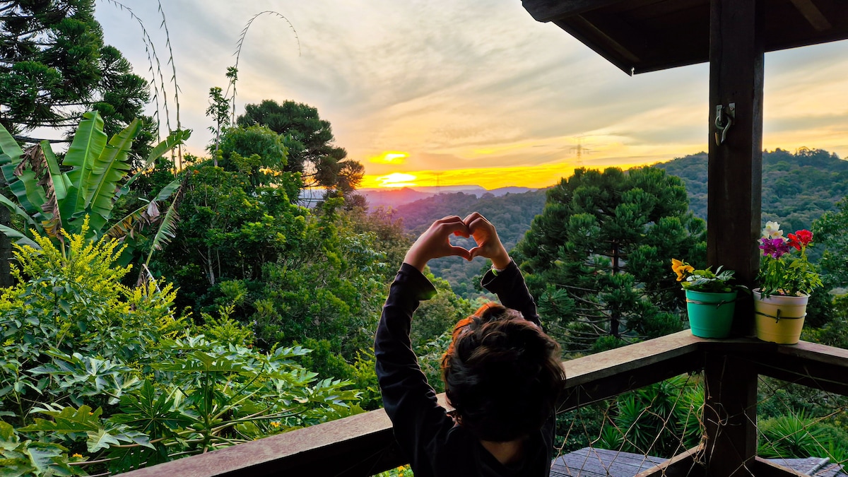 A child stands on a wooden deck, forming a heart shape with their hands against a vibrant sunset backdrop. Lush greenery and blooming potted flowers are visible, complementing the serene landscape of mountains and trees in the distance.