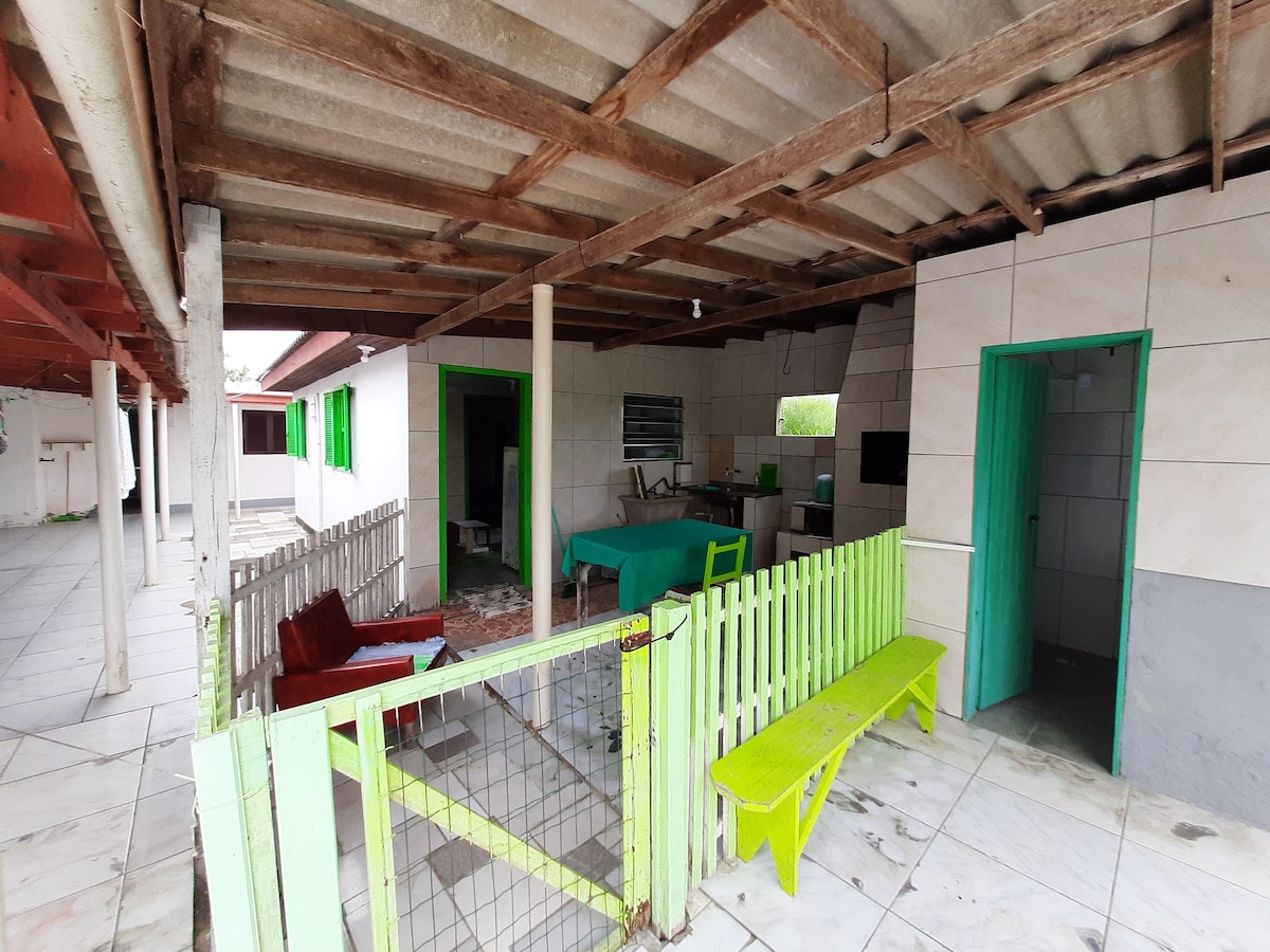 An outdoor area is enclosed with a green picket fence. Wooden furniture includes a small green bench, and a couple of simple seating options. The area is covered with a slanted roof, with light-colored walls and green doors visible, creating a relaxed, open gathering space.