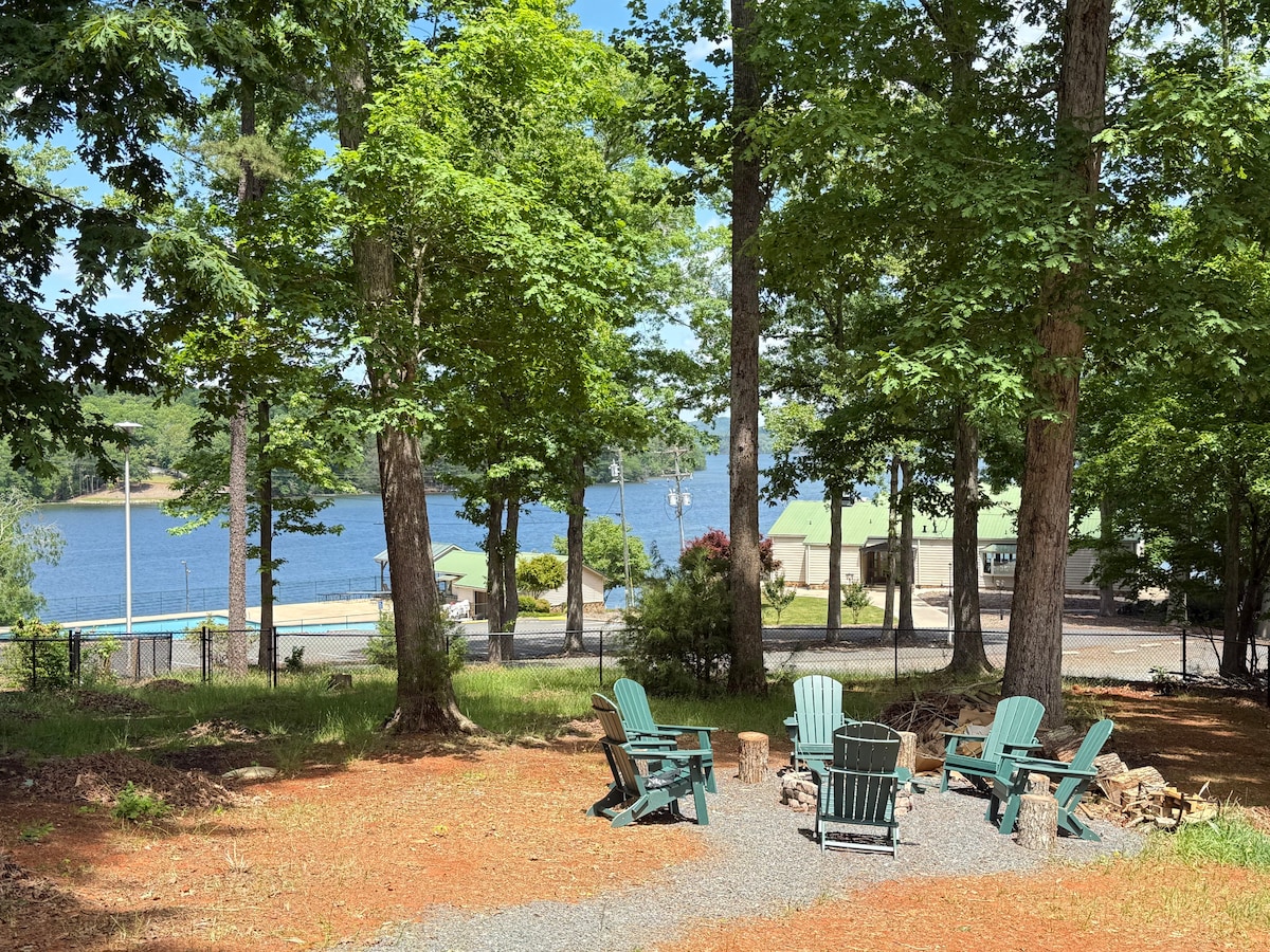 A serene outdoor space features several green Adirondack chairs arranged around a small fire pit. Tall trees provide shade, and a view of Lake Tillery is visible in the background, with a gentle shoreline and nearby structures peeking through the foliage.