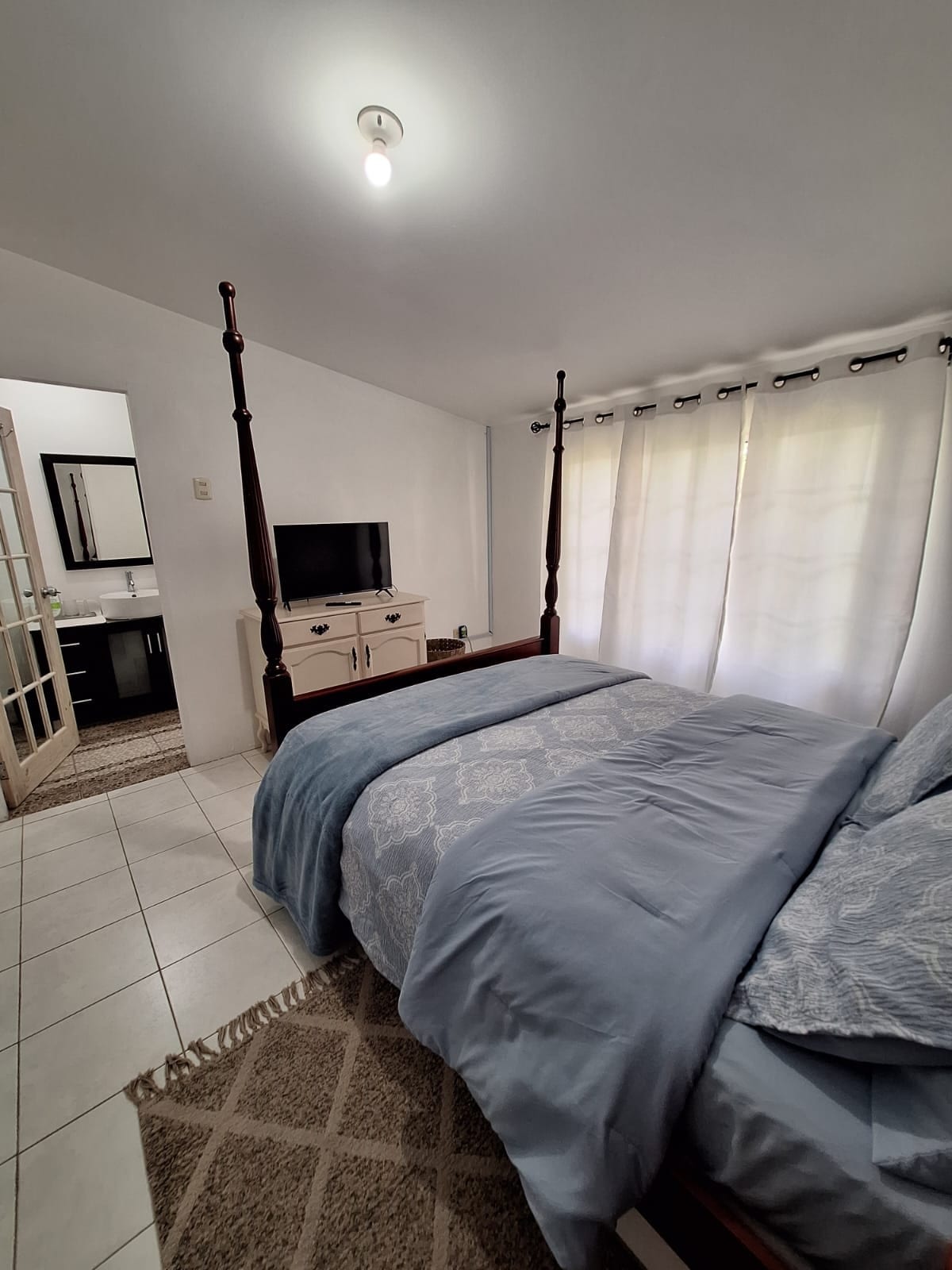 A cozy bedroom features a four-poster bed with soft blue bedding and a textured area rug. A television sits atop a light-colored dresser, and light flows in through sheer curtains. A bathroom doorway is visible in the background, offering convenient access.