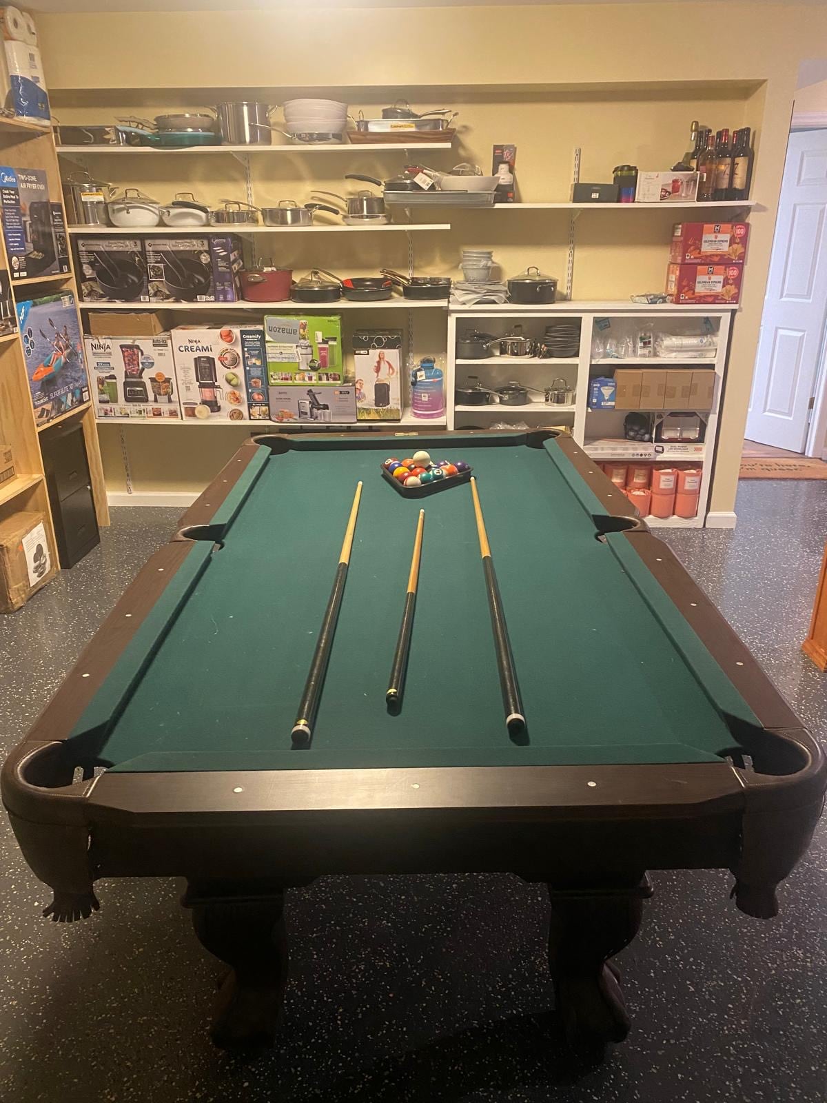 A billiard table is positioned in a well-organized room, featuring a green felt surface and three cues resting across the table. Shelves in the background display a variety of items, including appliances and storage containers.