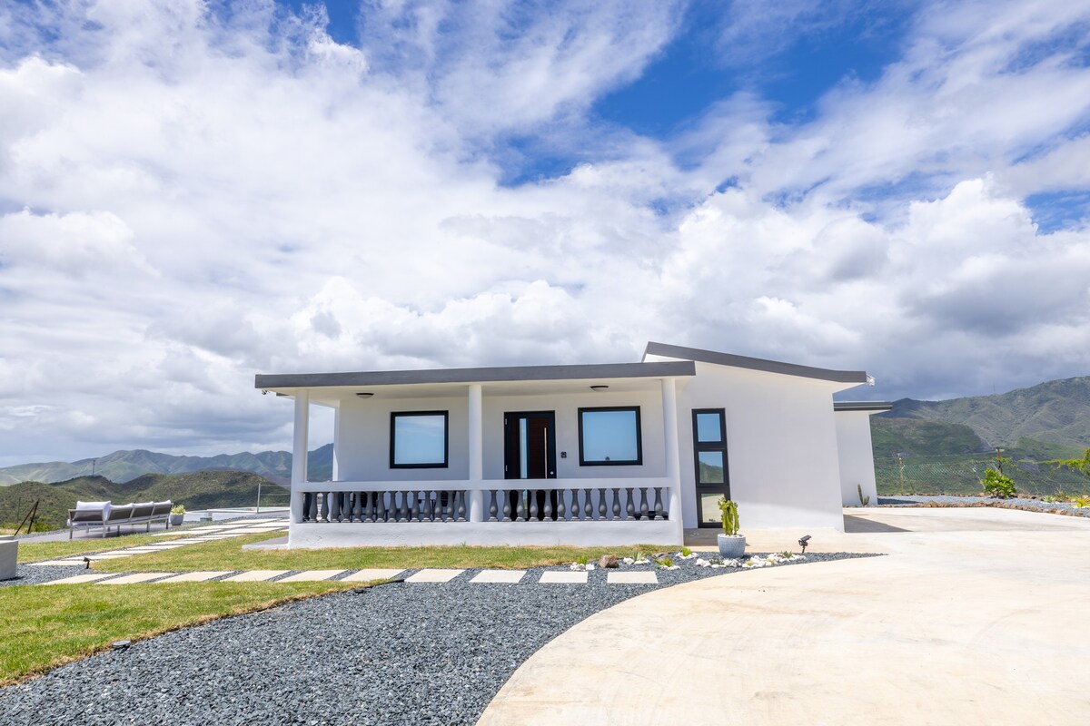 A modern one-story house is situated amidst a scenic landscape, featuring large windows and a covered porch. The pathway leads to a well-maintained lawn with decorative stones and potted plants. Expansive mountain views are visible in the background, under a partly cloudy sky.