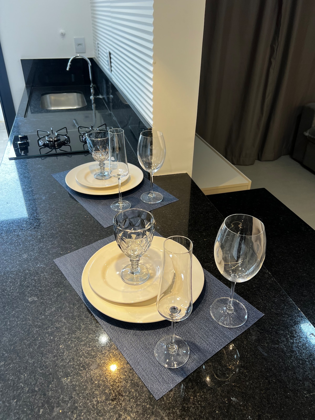 A sleek kitchen counter showcases three place settings, each featuring a plate, a decorative glass, and a tall stemmed glass. A gas stove is visible in the background, with light reflecting off the dark granite surface.