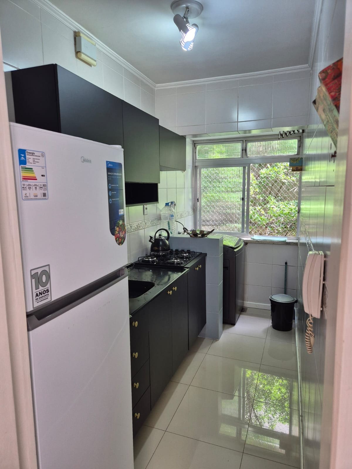 The kitchen is equipped with dark cabinetry, a white refrigerator, and a gas stove. A window allows natural light to enter, with views of greenery outside. Light tile flooring contributes to a clean and functional space.