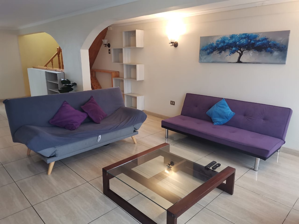 A living area is presented with two futons adorned with purple and blue cushions. A wooden table with a glass surface sits between them. Light walls and floor tiles create a bright atmosphere, complemented by a wall-mounted art piece featuring a blue tree.