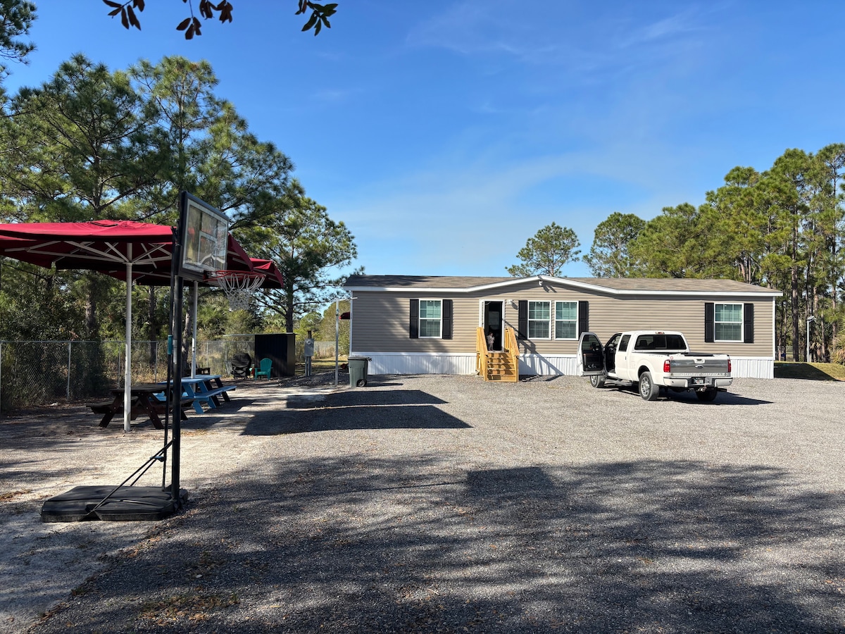 A spacious outdoor area features a gravel parking lot, accommodating vehicles and recreational equipment. A basketball hoop stands close to the home, which displays multiple windows. Surrounding trees offer shade, contributing to a serene atmosphere in this country setting.