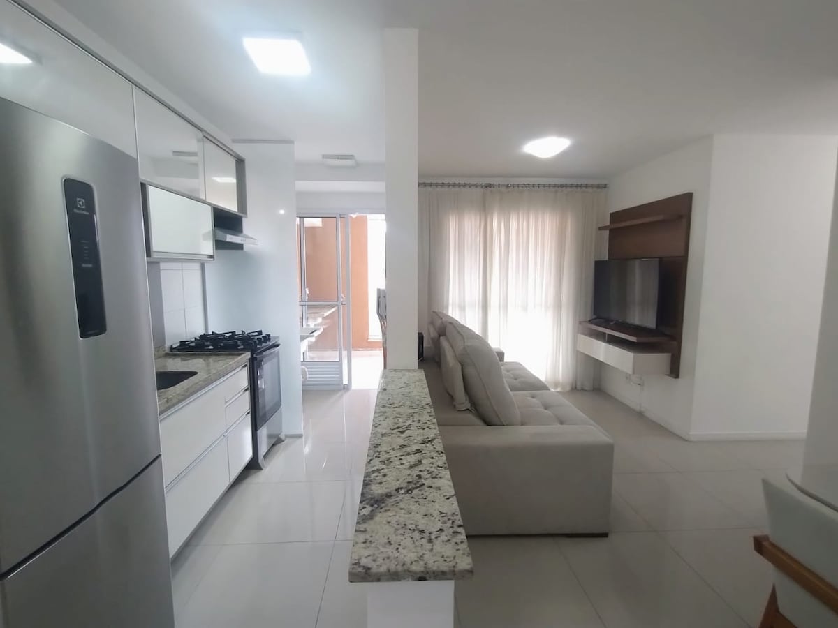 A modern kitchen space features sleek cabinetry and stainless steel appliances, complemented by a granite countertop. A comfortable seating area includes a light-colored sofa and a television mounted on a wooden panel, with large windows allowing natural light to fill the room.