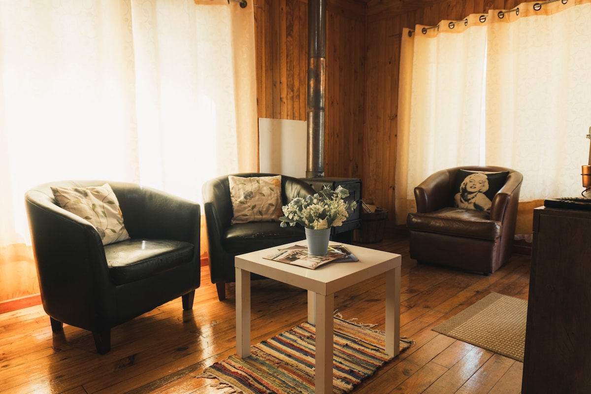 A cozy living area is furnished with two black armchairs and one brown leather chair, complemented by a small white coffee table at the center. Natural light is filtered through sheer curtains, enhancing the warm wooden walls and flooring.