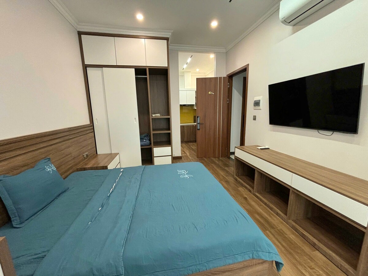 A modern bedroom features a spacious bed dressed in a blue cover. A large television is mounted on the wall opposite the bed. Storage cabinets and a dresser are situated nearby, with warm wood tones complementing the overall design. Large windows allow for natural light.