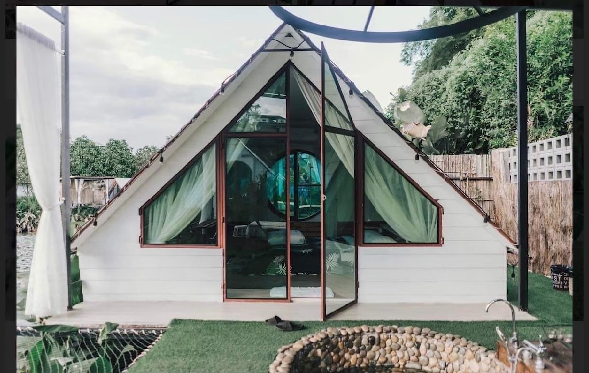 A unique a-frame structure features large, triangular windows providing natural light. The entrance is framed by flowing curtains, while a circular window adds a distinctive touch. The surrounding area includes greenery and a small stone-paved water feature, enhancing the serene environment.