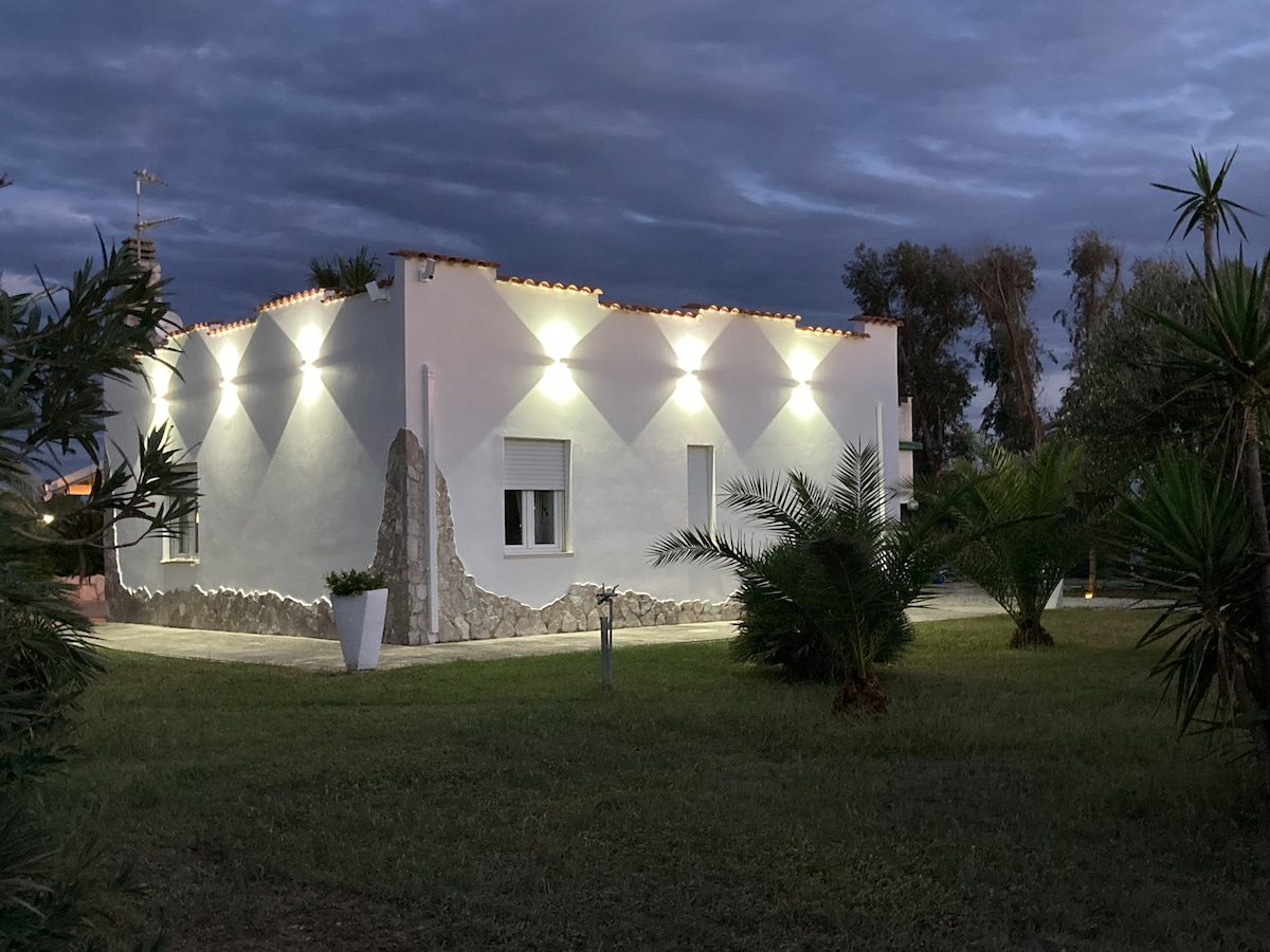 The white hacienda-style house is illuminated with soft exterior lighting, creating a serene ambiance against the evening sky. Palm trees and landscaped greenery frame the structure, highlighting its modern design and inviting entrance.