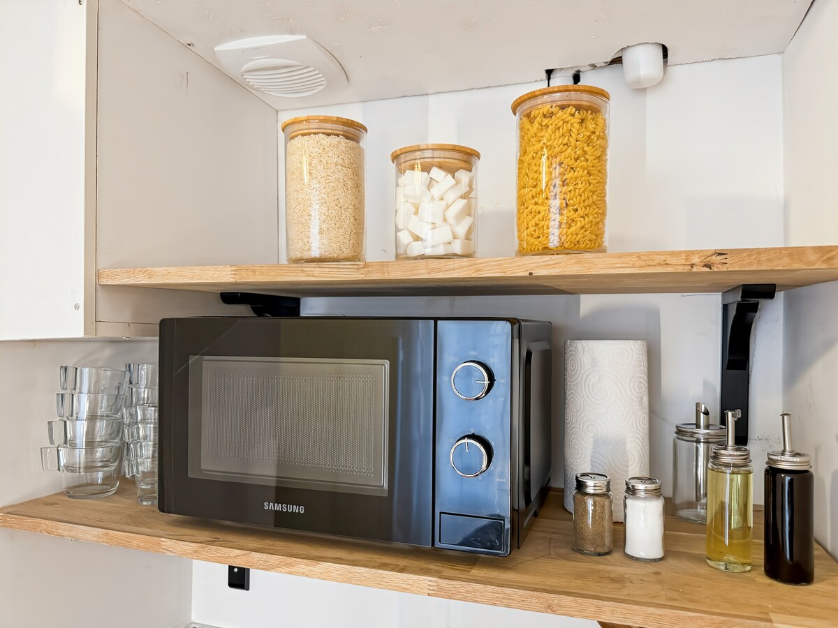 A microwave is positioned on a wooden shelf beneath glass jars containing pasta, sugar, and rice. Clear glasses and condiments, including salt, pepper, and oil, are neatly organized nearby, contributing to a functional kitchenette atmosphere.