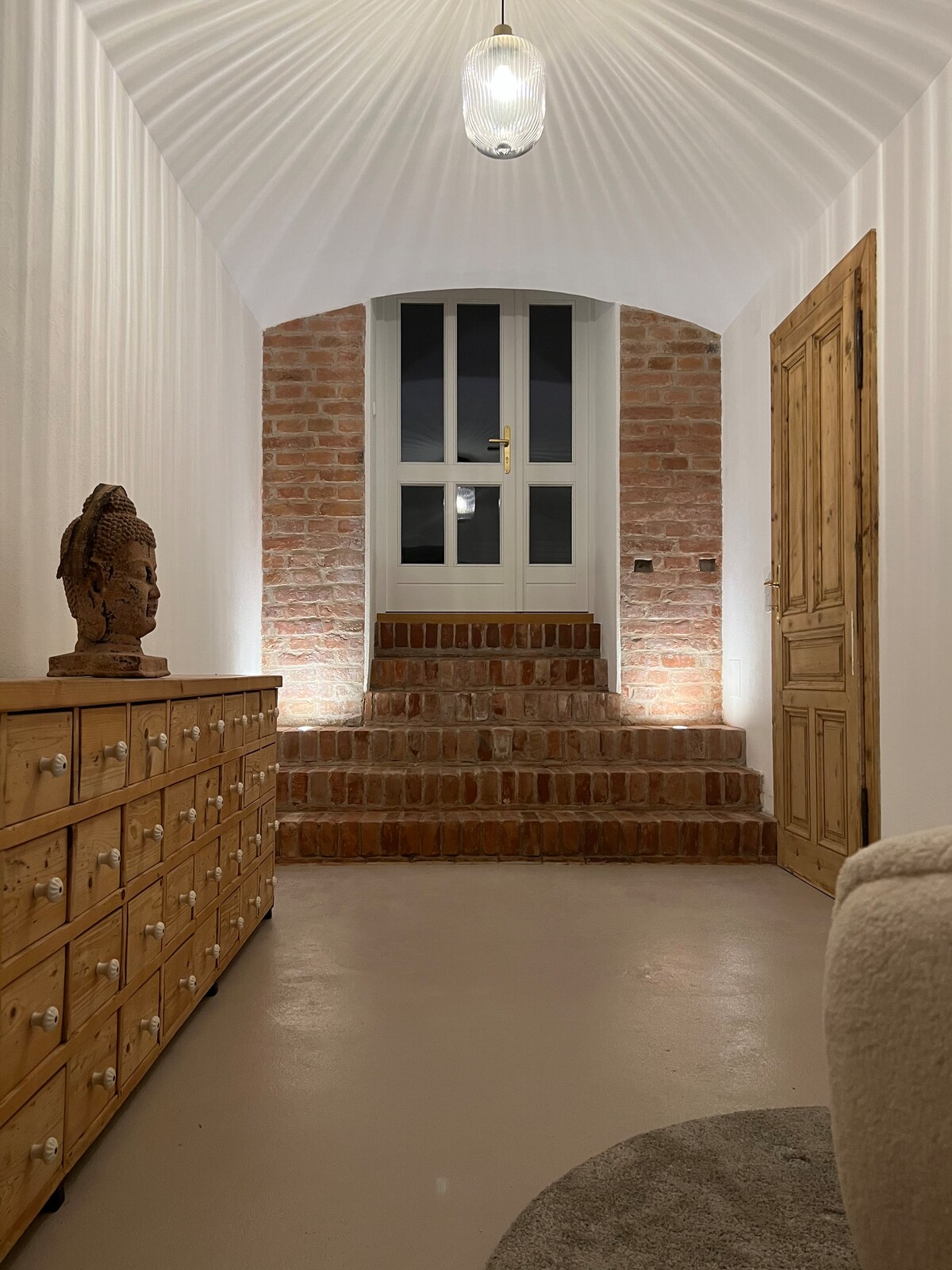 An entryway is presented with a warm wooden cabinet featuring multiple drawers. The brick walls offer a classic look, while a softly illuminated staircase leads to large glass doors. A welcoming light fixture hangs from the ceiling, enhancing the overall ambiance.