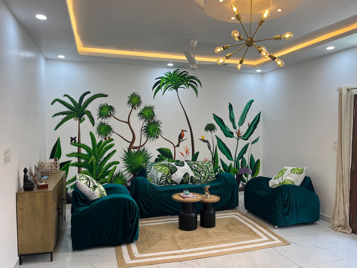 A living area is adorned with vibrant greenery-themed wall art depicting various plants and birds. Plush teal sofas, accented with decorative cushions, accompany a pair of dark coffee tables positioned over a textured rug. Soft lighting highlights the modern fixtures and brightens the inviting space.