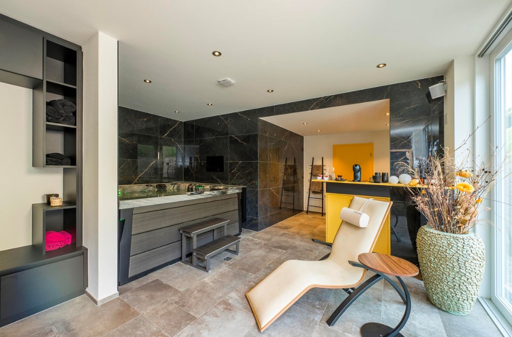 A modern spa area is showcased with dark marble walls and large windows. A sleek lounge chair is positioned near a stylish bathtub. A small wooden table complements the space, while a decorative vase filled with dried branches adds a natural touch. The room is illuminated by recessed lighting.