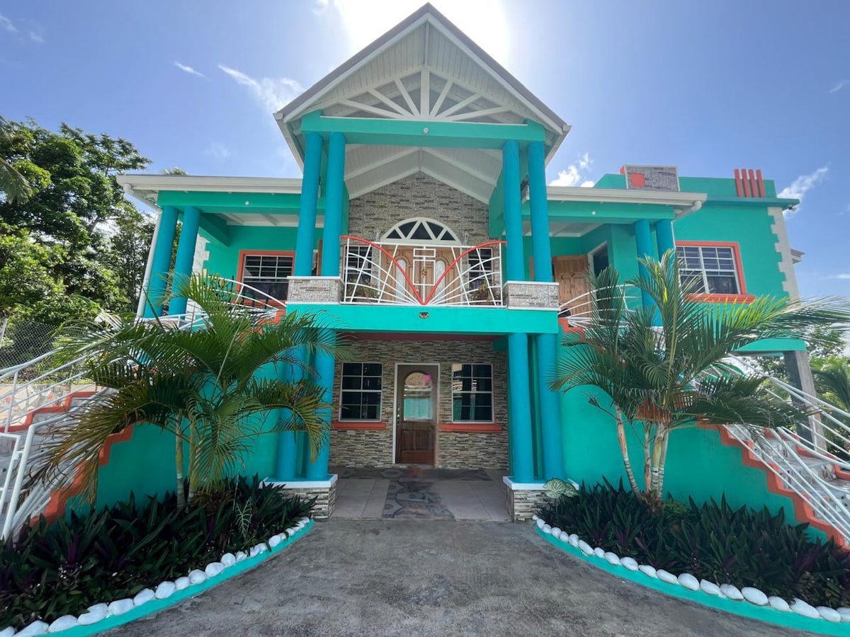 A vibrant two-story house features a striking turquoise exterior, complemented by white accents and lush greenery. The entrance is marked by bold columns and a spacious front porch, inviting guests into a welcoming retreat.