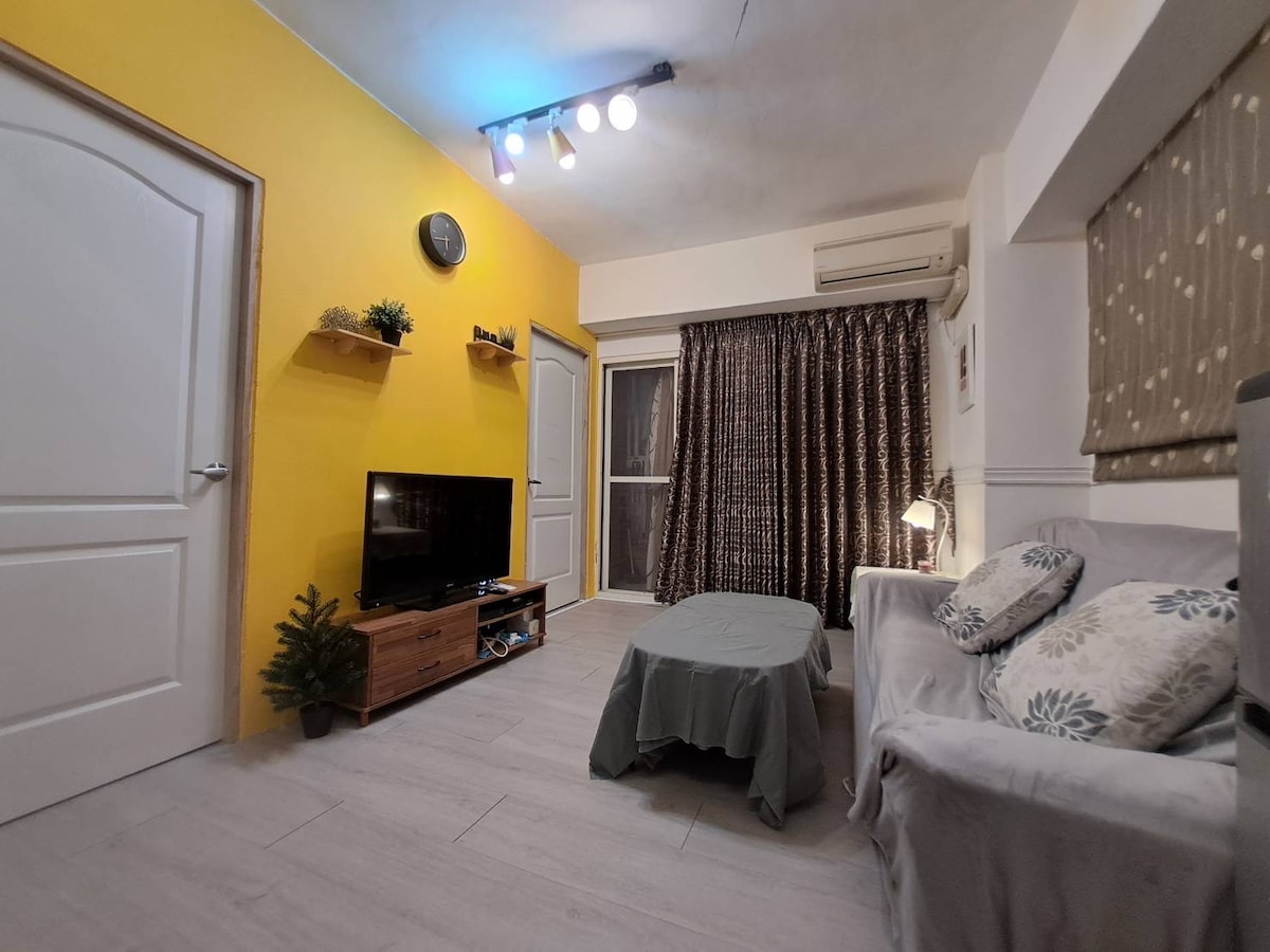 A living room features a yellow accent wall and wooden television stand. A gray sofa is positioned in front of a small table, with soft lighting from a lamp accentuating the area. Floor-to-ceiling curtains frame a window, and an air conditioning unit is visible.