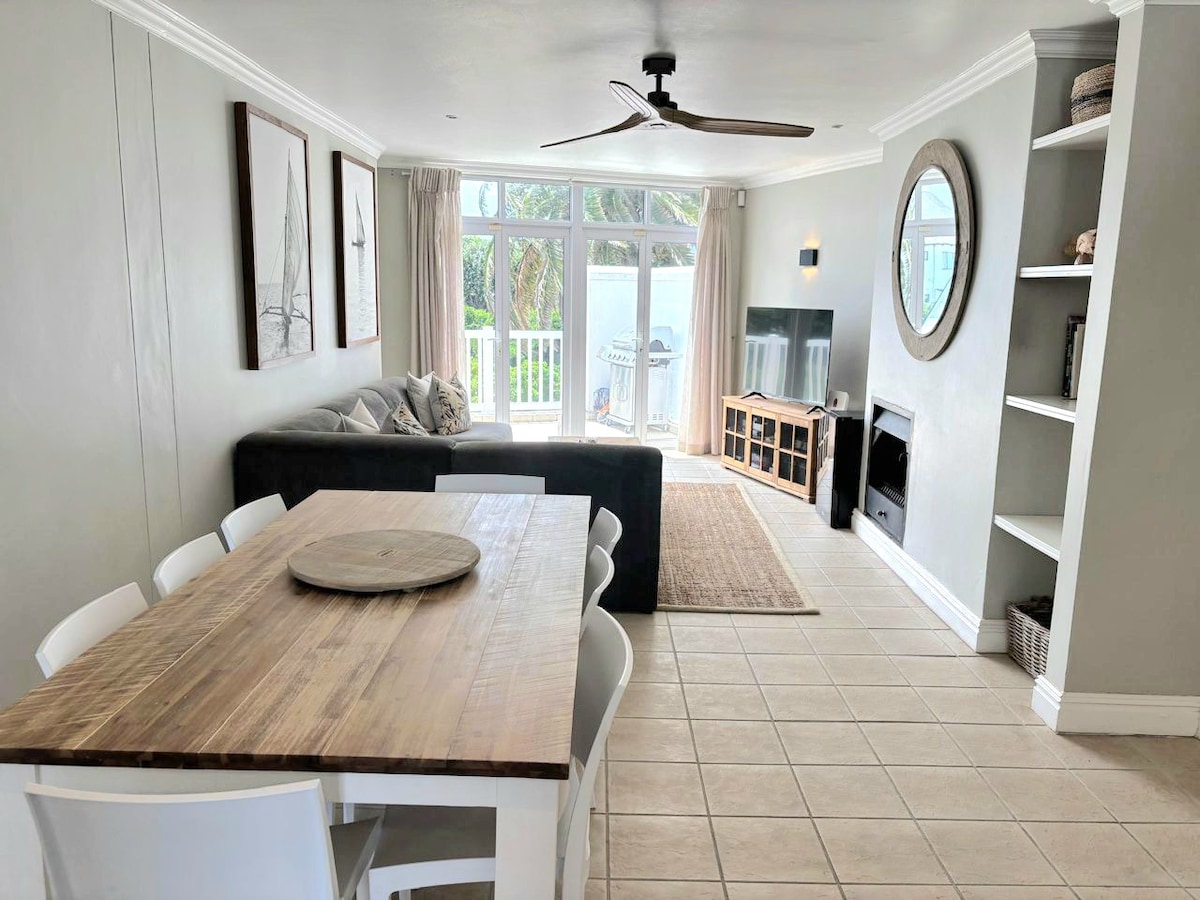 A spacious living area features a large dining table with six white chairs, a comfortable dark sofa, and a flat-screen TV mounted on the wall. Natural light streams in through glass doors that lead to an outdoor area, while decorative elements provide a calming ambiance.