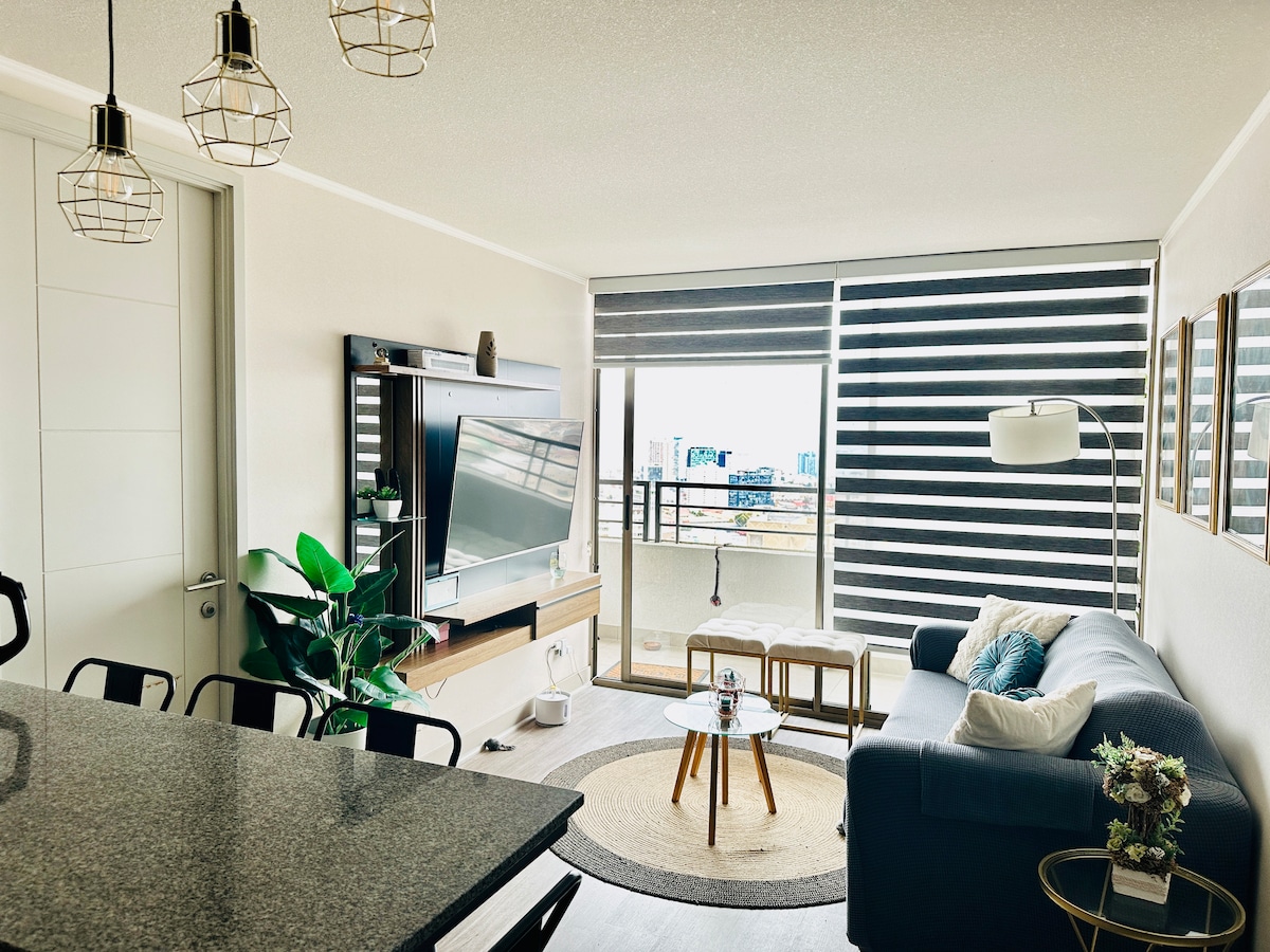 A modern living area is showcased, featuring a dark sofa complemented by a light rug. A television is mounted on a wooden panel, and large windows with striped blinds provide an urban view. Potted plants add a touch of greenery, enhancing the inviting atmosphere.