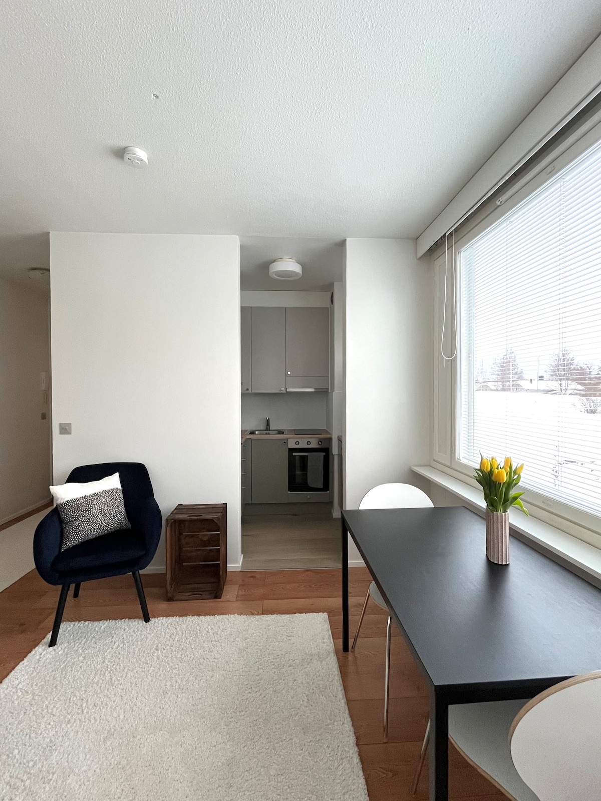 A modern living space is depicted, featuring a small dining table with two chairs and a cozy armchair. A well-equipped kitchen is visible in the background. Natural light filters through large windows, illuminating the minimalist decor and a vase of tulips on the table.