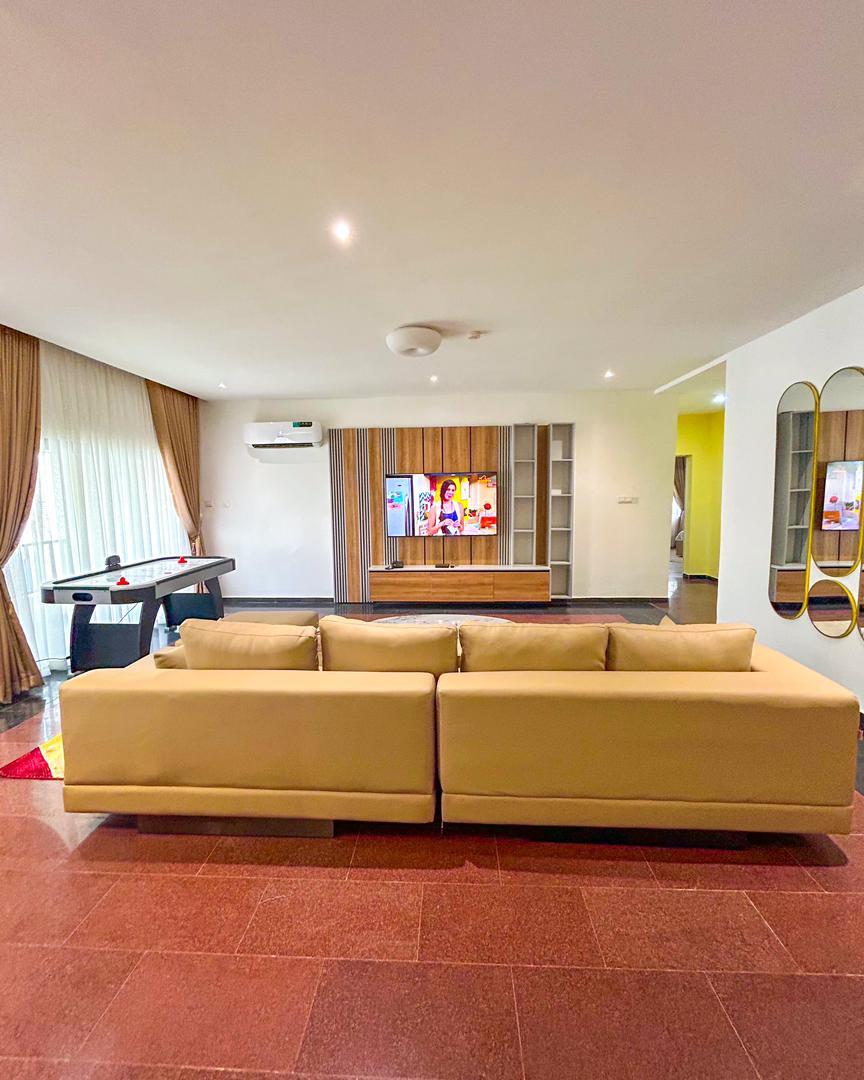 A light-filled living area features a neutral-colored sofa facing an entertainment unit with a television. A game table is visible to the left, while large windows with sheer curtains allow natural light to illuminate the space. Decorative mirrors add depth to the room.