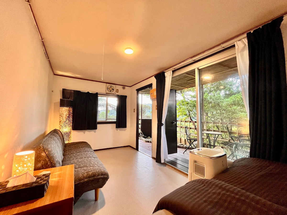 A cozy room is equipped with a comfortable seating area featuring a patterned sofa. Large glass doors offer access to a balcony with outdoor seating, allowing for natural light to brighten the space. Soft lighting creates a relaxing ambiance.