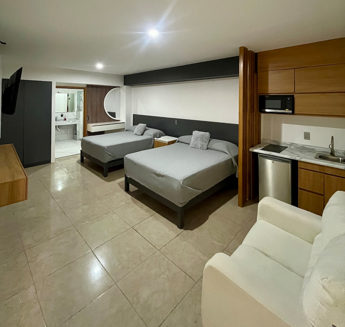 A spacious room features two large beds with neutral bedding arranged on dark wooden frames. A kitchenette with a small refrigerator and microwave is visible. A comfortable seating area is provided with a light sofa, enhancing the modern design. Soft lighting is present throughout the area.