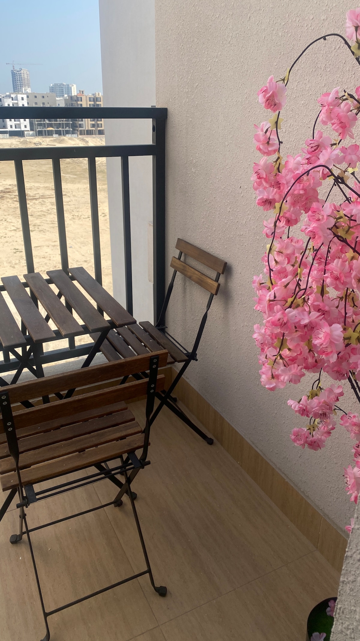 A small balcony features a wooden folding table accompanied by two matching chairs, set against a neutral wall. Delicate pink flowers in a pot add a touch of nature, enhancing the serene outdoor space. The view includes a distant urban landscape.